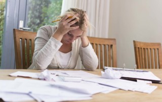 Frustrated, caucasian woman sitting at a table with her head in her hands, looking at papers spread all over table.