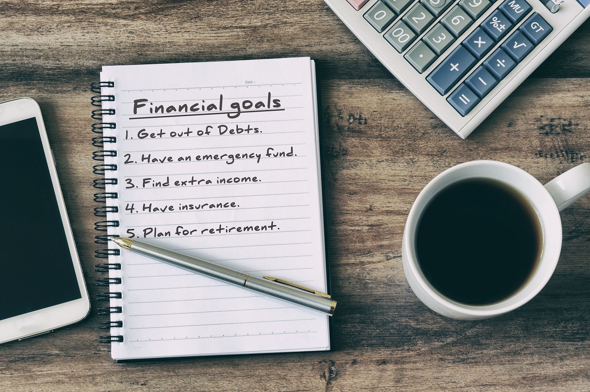 Aerial view of a notebook with financial goals written on it, a coffee cup, calculator, and pen