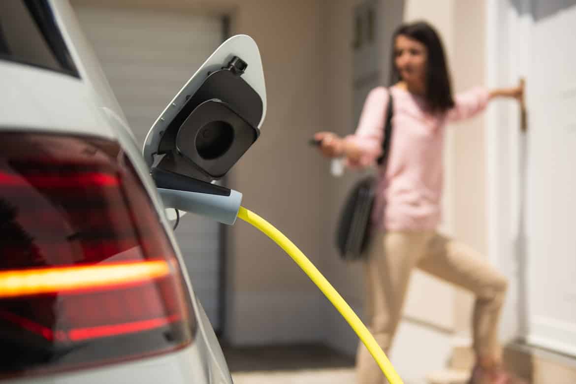 An electric vehicle plugged into a power outlet with a young woman blurred in the background locking the car