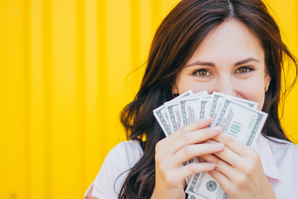 Brunette woman smiling holding money in front of her face, fanned out.