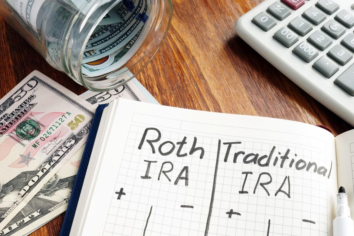 An open journal on a table with the words Roth IRA and Traditional IRA written in marker at the top. Calculator and money in the background.