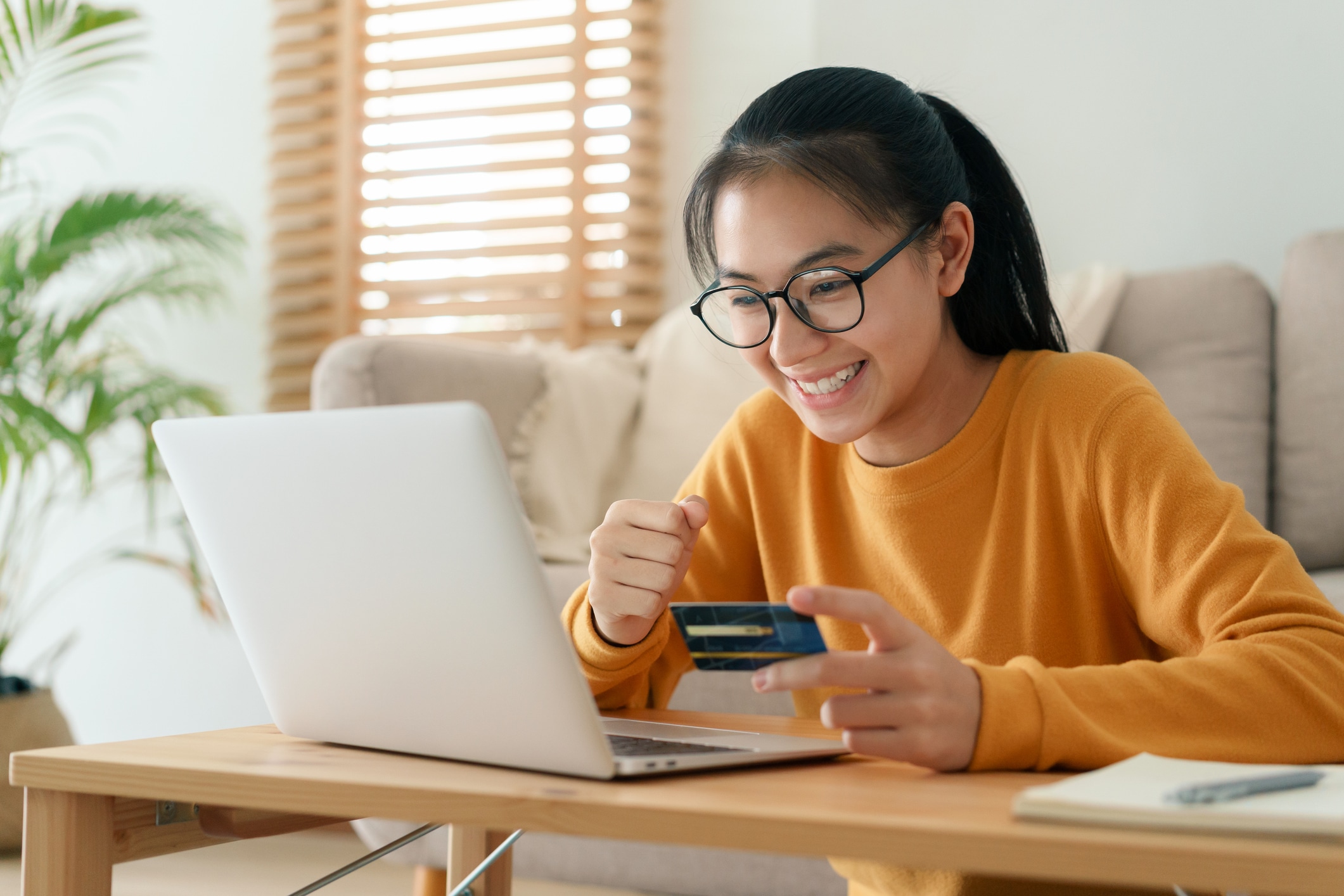 Smiling person wearing glasses sits at a table using a laptop while holding a credit card, appearing to shop or make a payment online in a bright living room