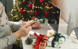A person wearing a cozy sweater shops online at a laptop near a decorated Christmas tree, with wrapped holiday gifts and ornaments arranged on the desk.