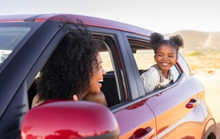 A smiling woman in the driver’s seat looks back at a young child leaning out of the rear passenger window of a red car, sharing a joyful moment during a sunny drive