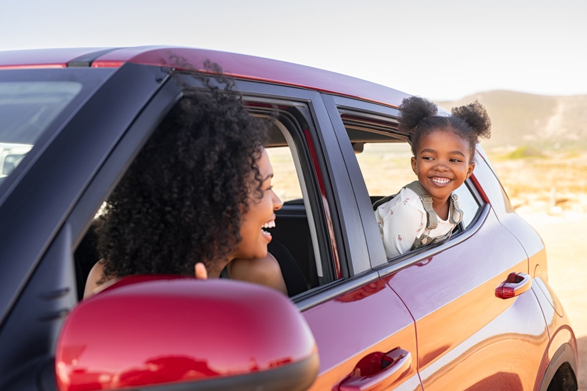 A smiling woman in the driver’s seat looks back at a young child leaning out of the rear passenger window of a red car, sharing a joyful moment during a sunny drive